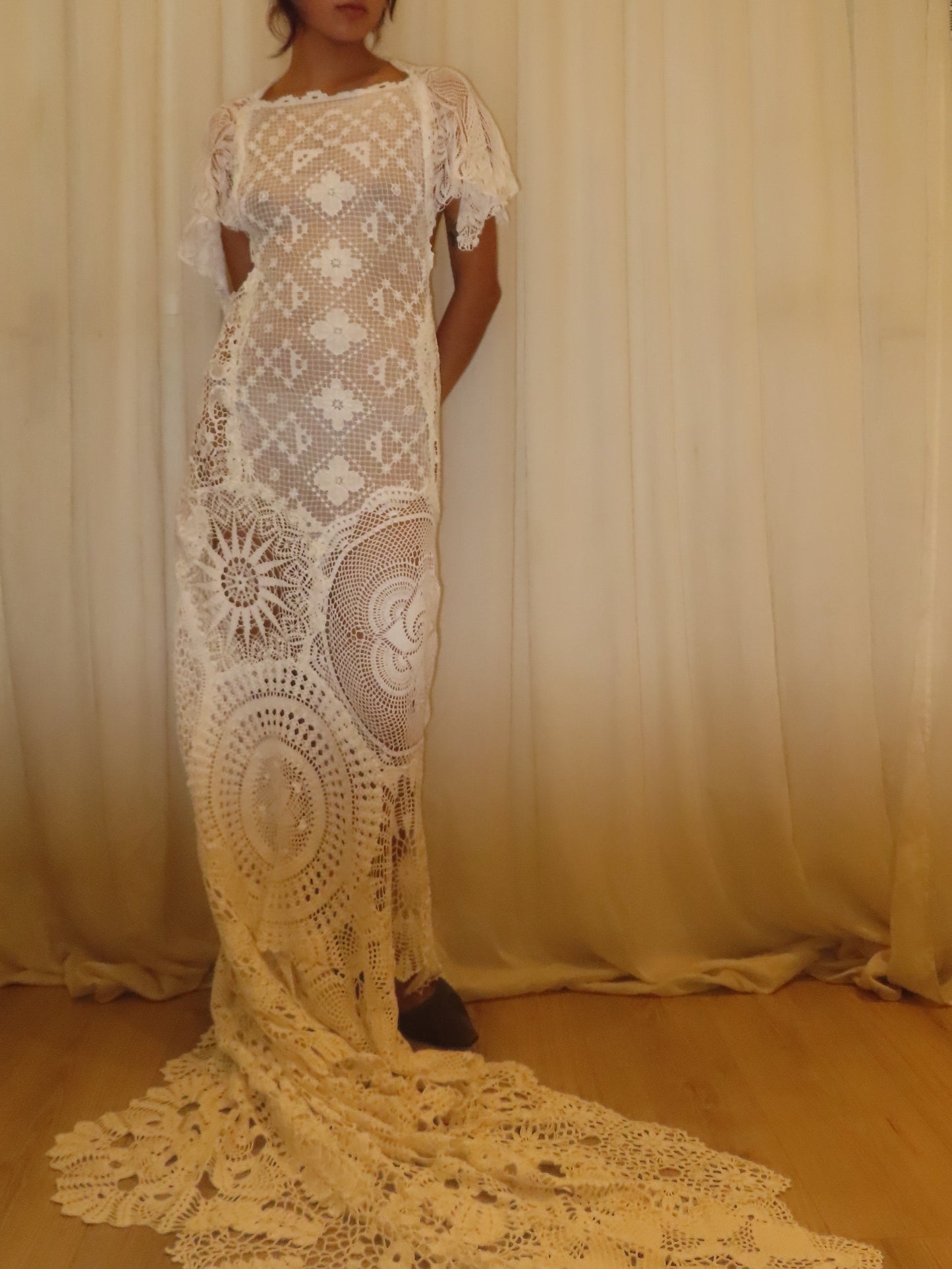 A woman wearing a cream-colored crochet lace dress with short sleeves and a long train, standing in front of a draped cream fabric backdrop in an indoor room with wooden floors.