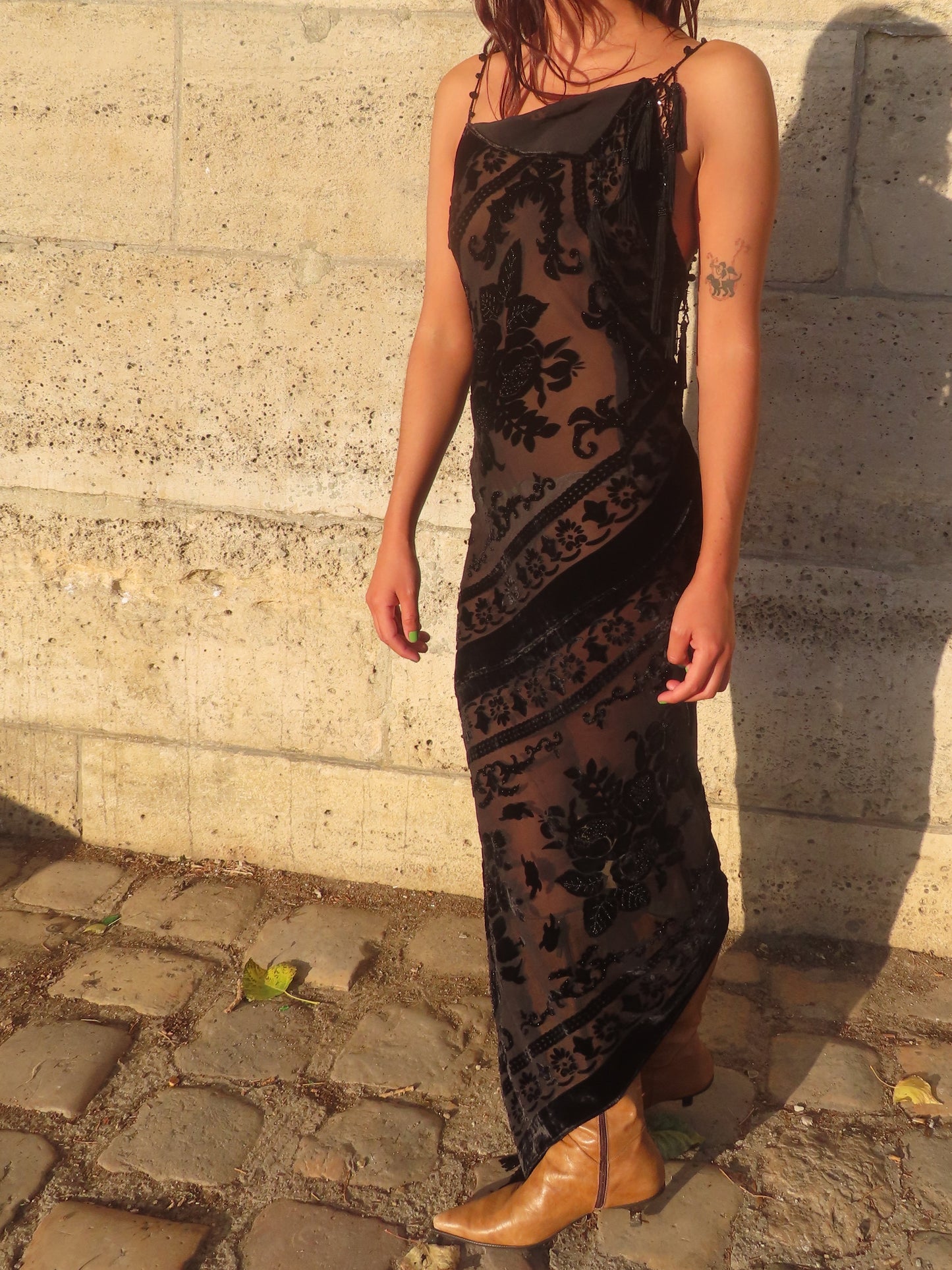 A woman wearing a black sheer floral lace maxi dress and tan boots, standing outdoors against a light stone wall with her shadow cast on the background.