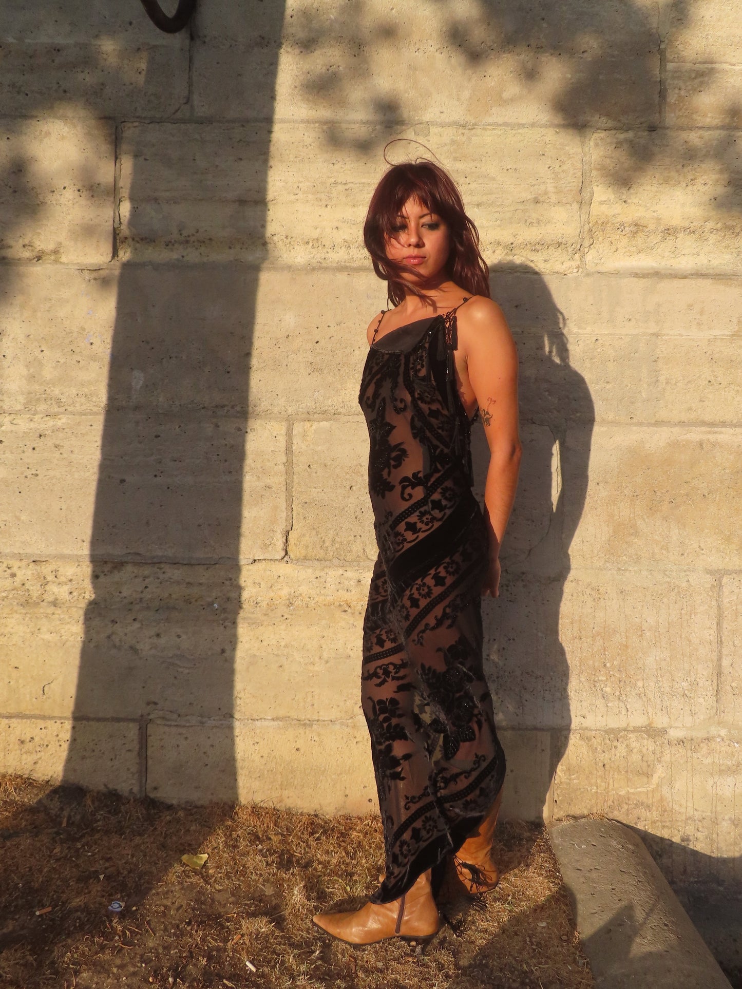 A woman wearing a black velvet bias dress with a bohemian pattern, standing against a sunlit concrete wall in Paris
