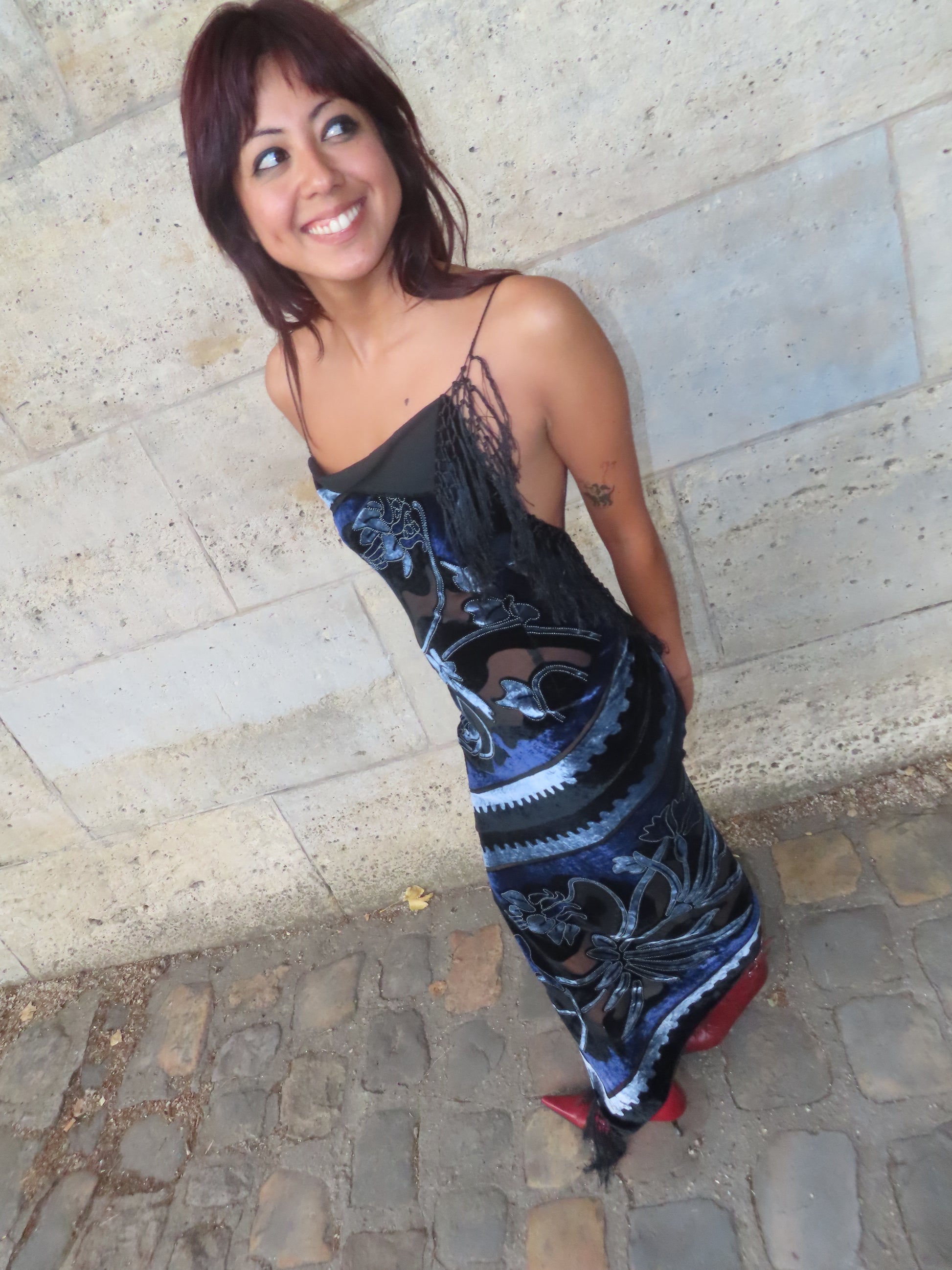 A woman wearing a black spaghetti-strap maxi dress with a blue and black abstract pattern, standing on a cobblestone street against a stone wall.