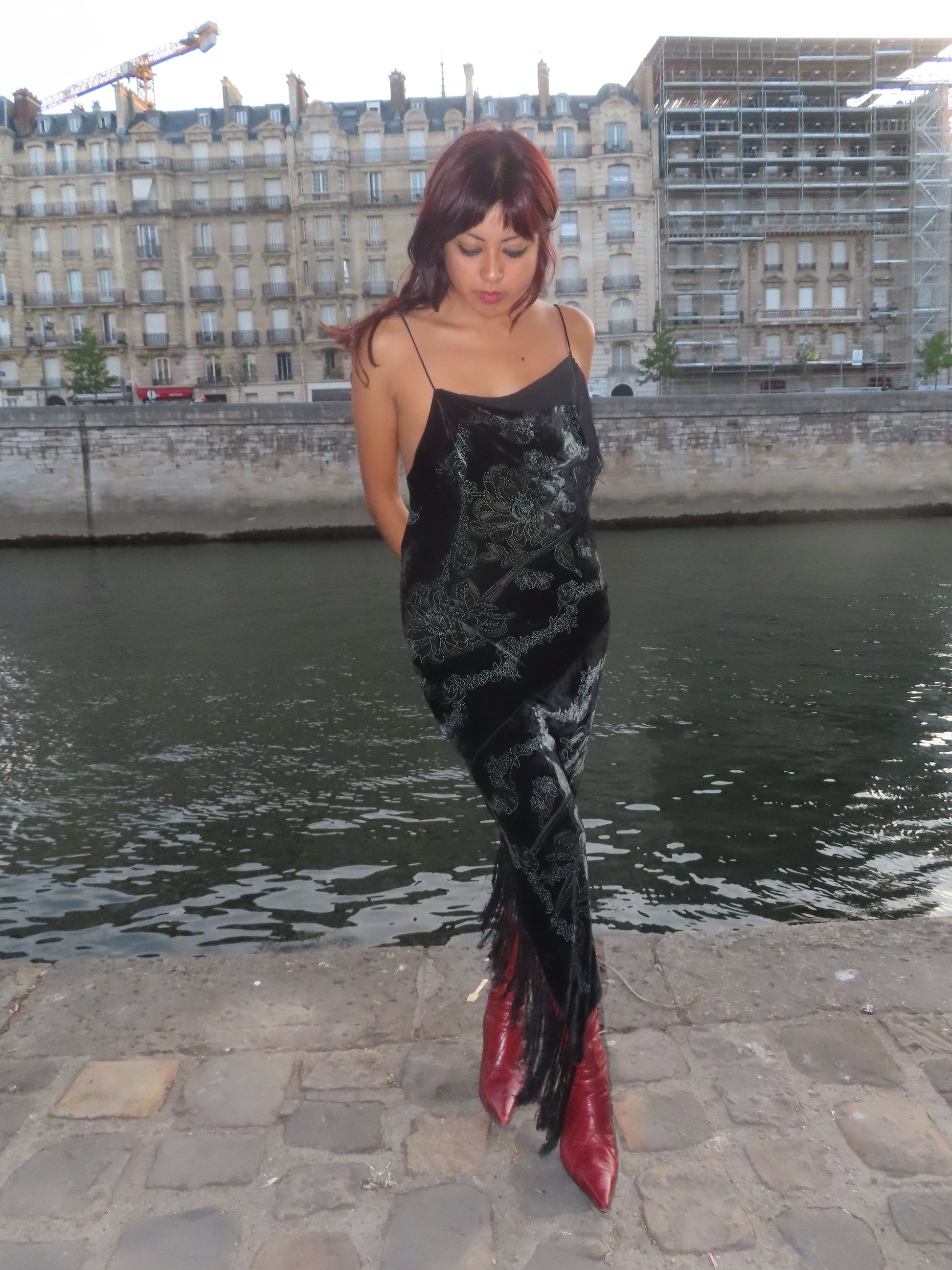 A woman stands on a stone quay beside a river with Parisian-style buildings in the background. She is wearing a black sleeveless dress with a dark floral pattern and fringe at the hem, paired with red pointed-toe boots.