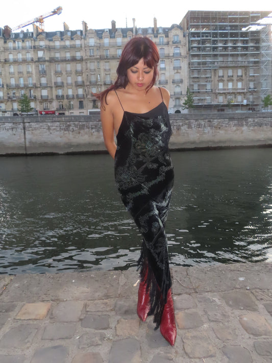 A woman stands on a stone quay beside a river with Parisian-style buildings in the background. She is wearing a black sleeveless dress with a dark floral pattern and fringe at the hem, paired with red pointed-toe boots.