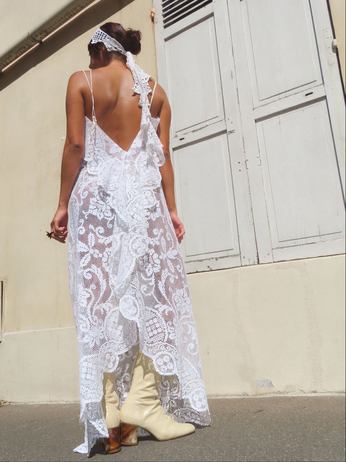 A woman stands outdoors with her back to the camera wearing a white lace backless dress with thin straps, cream boots, and a white lace headband, in front of a beige building wall with white shutters.