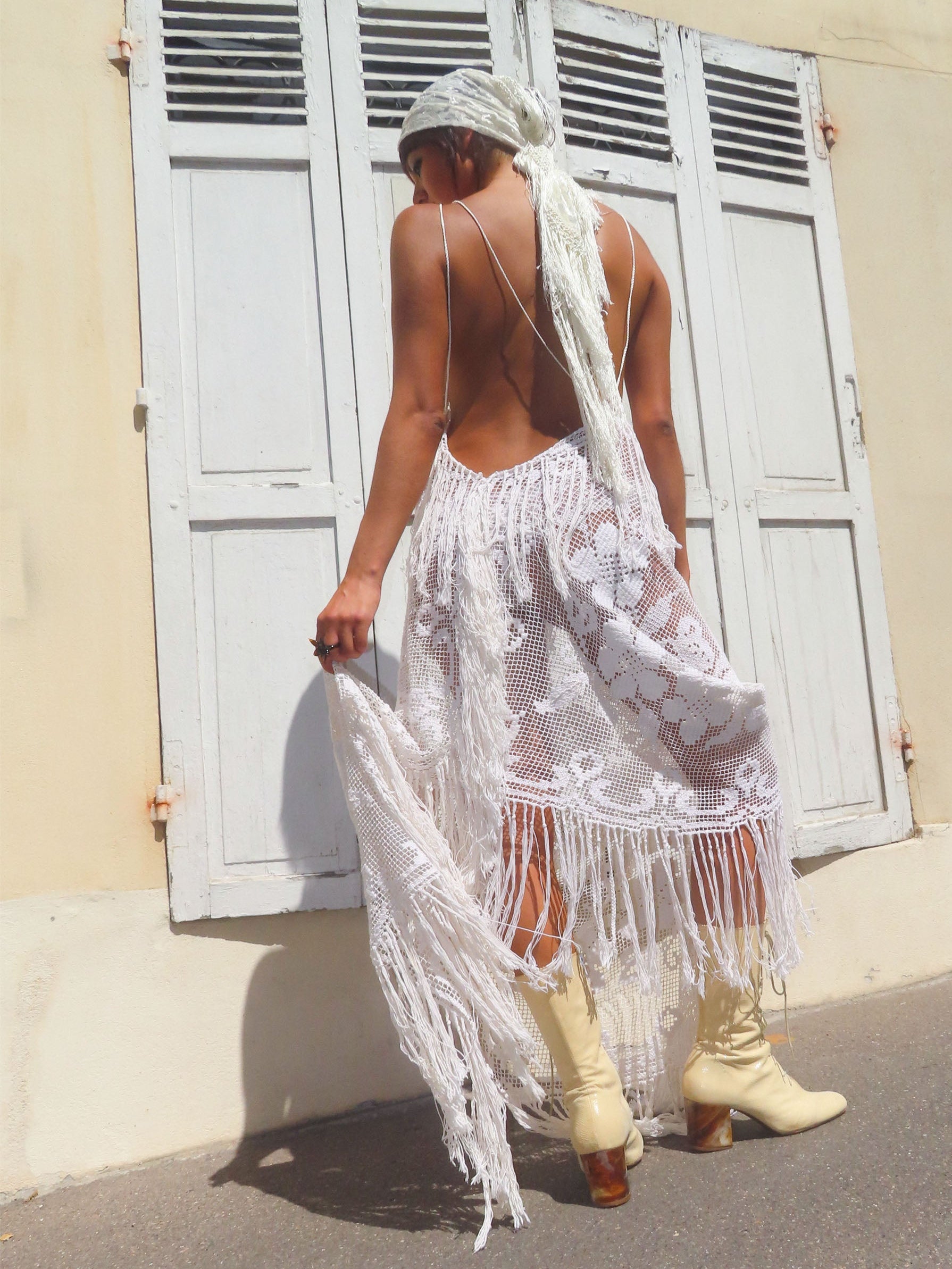 A woman stands outdoors in front of a beige wall with white shutters, photographed from behind. She is wearing a white lace boho-style skirt with long fringe, a matching white headscarf, and light beige heeled boots.
