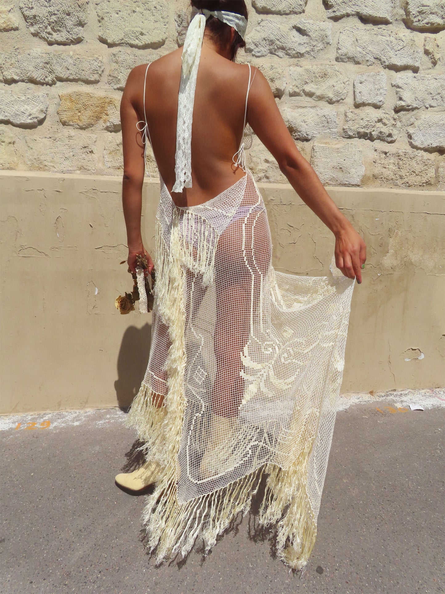 A woman shown from behind wearing a long, open-back crochet cover-up dress in cream/off-white with fringe and mesh details, standing outdoors against a stone wall.
