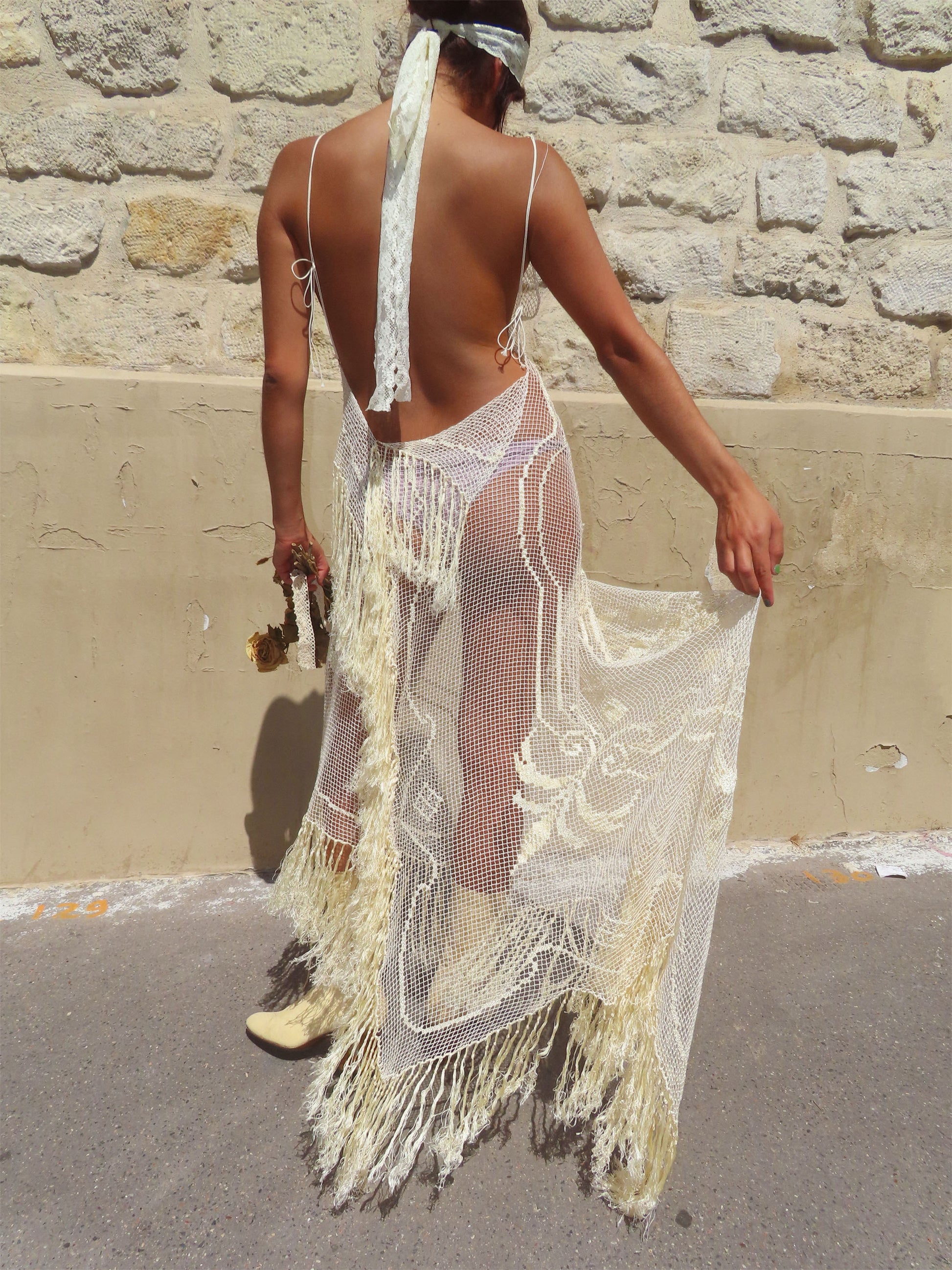 A woman shown from behind wearing a long, open-back crochet cover-up dress in cream/off-white with fringe and mesh details, standing outdoors against a stone wall.